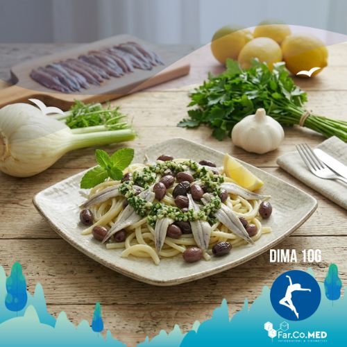 Primo piano fotografico in formato 1:1 di una insalata di alici marinate condita con erbe aromatiche e olive, impiattata scenograficamente. Sullo sfondo, leggermente sfocati per dare profondità, si distinguono un tagliere con alici, limoni freschi, un mazzo di prezzemolo, un aglio e un finocchio, il tutto adagiato su una superficie rustica in legno.