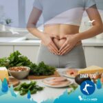 Una donna che fa il simbolo del cuore con le mani, all'altezza dell'ombelico per indicare il microbiota collegato alla dieta chetogenica e intestino. Davanti a lei ci sono anche una varietà di alimenti chetogenici come formaggi, pesci e verdure a foglie verdi
