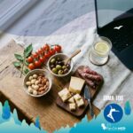 Un'inquadratura dall'alto di una varietà di spuntini dietetici fuori casa, con noci miste, olive verdi e nere, cubetti di formaggio parmigiano con un piccolo coltello da formaggio e bastoncini di mini salame su un tagliere di legno rustico e un panno di lino grigio chiaro. In secondo piano si vedono un bicchiere di acqua frizzante con una fetta di limone e un computer portatile, posizionati su un tavolo di legno vicino a una finestra. Il tutto è illuminato da luce naturale.