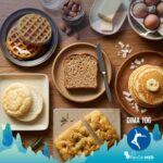 Una vista dall'alto di una ricca e invitante colazione senza carboidrati, ideale per la dieta chetogenica, disposta su un tavolo di legno rustico. L'immagine mostra cinque diverse alternative al pane: fette di mug bread tostato, chaffles croccanti, soffice cloud bread, una porzione di focaccia keto al rosmarino e pancake alla farina di cocco. Sono visibili anche ingredienti base come uova, burro, mandorle e rosmarino fresco.