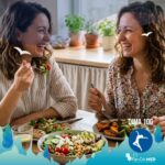 Due donne sorridono e chiacchierano felicemente sedute a un tavolo di legno in una cucina luminosa, gustando un pasto sano composto da insalate colorate, avocado, ceci e verdure grigliate. L'immagine trasmette un senso di benessere e convivialità, illustrando perfettamente il concetto di dieta del buon umore attraverso cibo nutriente e interazione umana autentica.